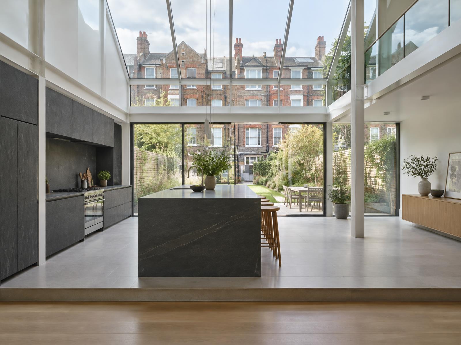 Double-height kitchen extension with a white steel frame, glass roof and dark stone island, looking onto Victorian terraces