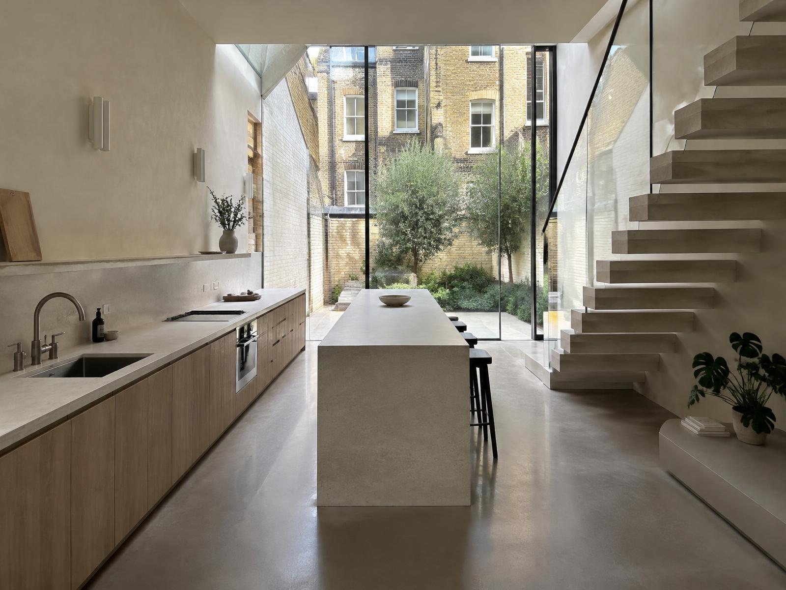 Narrow lower-ground kitchen with a floating stone stair and glazed rear opening onto yellow stock brick Marylebone backs